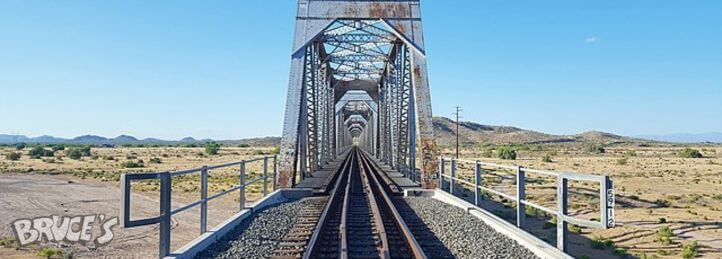 Railroad tracks running through a steel truss bridge over a desert landscape under a clear blue sky.