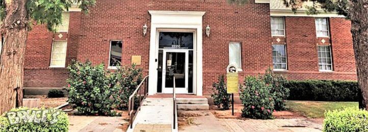 Brick building entrance with white trim, glass double doors, and shrubs lining a short walkway and ramp.
