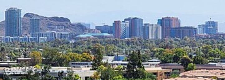 City skyline with modern high-rise buildings rising above a tree-filled neighborhood and distant mountains.